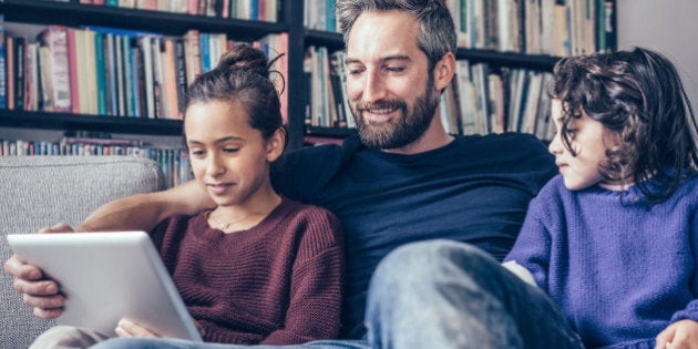 little family with bearded father and two daughters on sofa looking at tablet computer