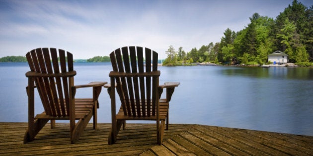 A couple of wooden Muskoka chairs sitting on the dock with a lake and cottages across in the background. Perfect for cottage related applications