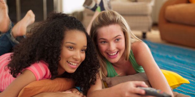 Two teenage girls lying down watching television with remote control smiling