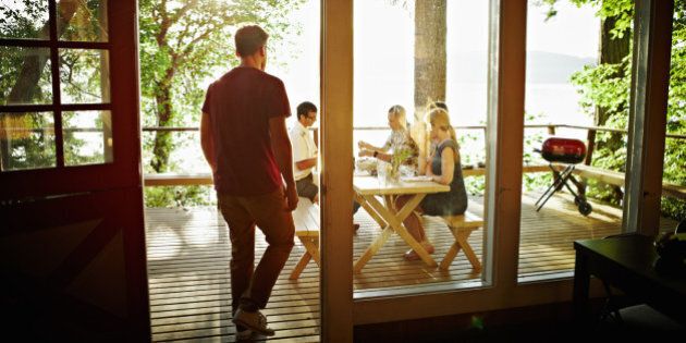 Man walking out door of rustic cabin to friends sharing a meal on deck at sunset