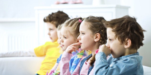 Side view on five cute kids sitting on sofa in domestic room and watching TV with interest. Focus on the girl keeping her finger in mouth.