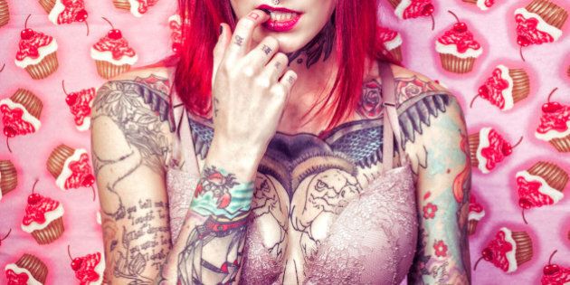 Sexy tattooed woman for valentine's day. Red hair, tattoos on a pink background with cupcakes and wearing devil horns.