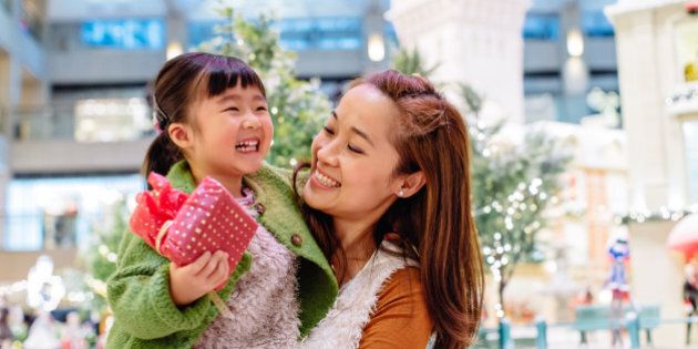 Pretty young mom carrying lovely little daughter who's holding a beautifully wrapped present in her hand, they both smiling joyfully while talking to each other in front of the lighting decoration in the shopping mall