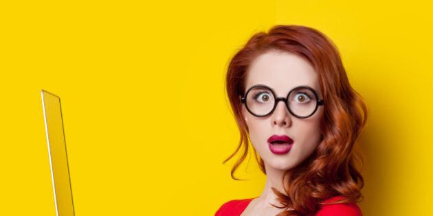 Surprised redhead girl with laptop computer in red dress on yellow background.
