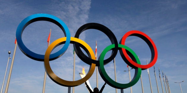 A spectator poses for a photo under the Olympic Rings at the 2014 Winter Olympics, Wednesday, Feb. 12, 2014, in Sochi, Russia. The Winter Olympics are in full swing, but warm temperatures in the Black Sea resort town of Sochi made heavy coats and scarves unnecessary on Wednesday. (AP Photo/Mark Baker)