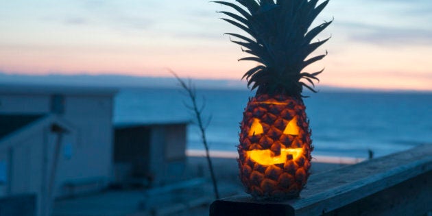 Jack o lantern halloween face carved into pineapple instead of pumpkin. Tropical fruit with ocean in background