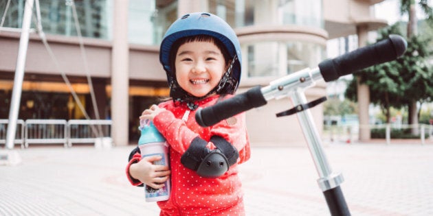 Lovely little girl with a helmet and protective gears smiling joyfully at the camera while holding her water bottle in front of the scooter on the street