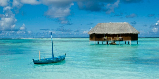 A dhoni (traditional Maldivian boat) floating in a lagoon with an over-water honeymoon bungalow off in the distance, Maldives.