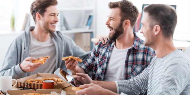 Three happy young men eating pizza together while sitting in the office