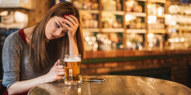 Pensive lonely young woman holding a glass of beer while sitting in a pub. She is looking at mobile phone and waiting for a call.