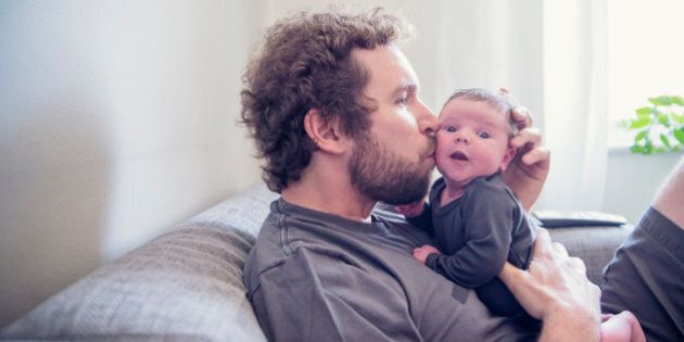 Young bearded dad kissing his 10 days newborn baby while relaxing on a couch at home. Both are wearing grey tops. Baby is very alert and have open eyes. TV remote control and window in background. Horizontal backlit waist up shot with copy space.
