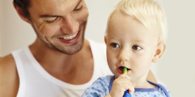 A little boy brushing his teeth while sitting on his dad's lap