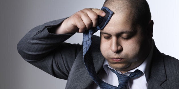 A hefty man in a suit wipes the sweat off his forehead with his tie. Photographed against a white background.