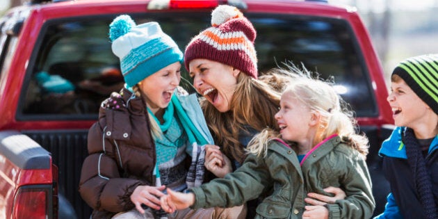 A happy mother with her three children outdoors in the fall, sitting on the back of a pickup truck, dressed for cool weather in jackets and winter hats. They are laughing together, having a good time.