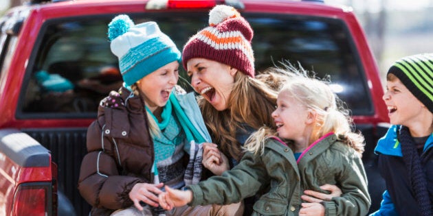 A happy mother with her three children outdoors in the fall, sitting on the back of a pickup truck, dressed for cool weather in jackets and winter hats. They are laughing together, having a good time.