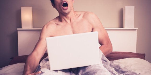 Young naked man is sitting in a bed and watching pornography on his laptop