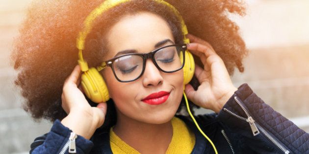 Attractive young woman listening music. Yellow headphones on head, eyeglasses, wear black jacket, yellow blouse. Hands on headphones, eyes closed.