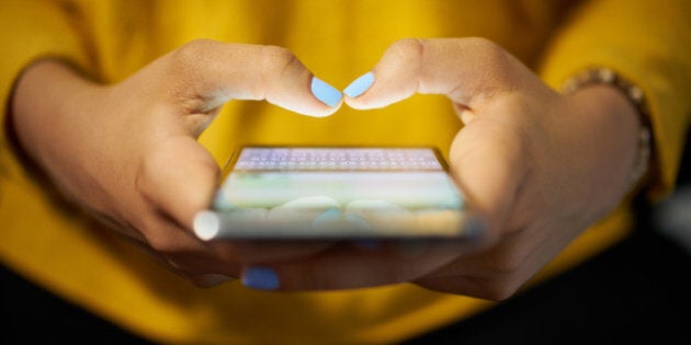 Young woman using cell phone to send text message on social network at night. Closeup of hands with computer laptop in background