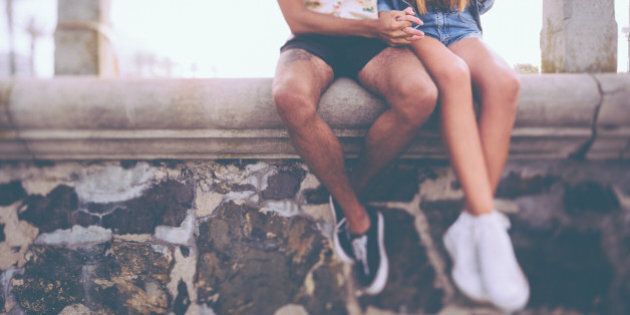 Cropped image of the legs of a hipster couple holding hands and sitting on a wall made of rough textured stone