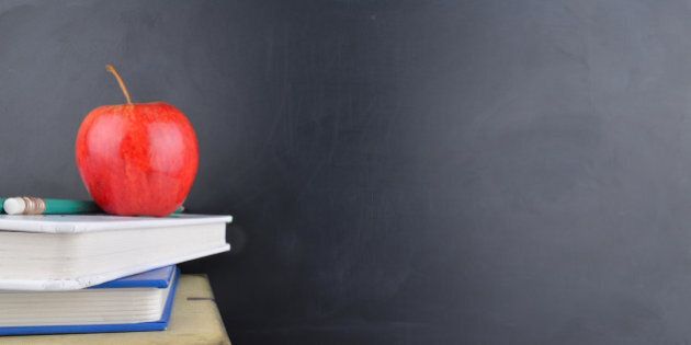 A classroom with a red apple, books and a blackboard with handwriting in white chalk on the board.