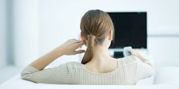 Young woman sitting in armchair watching TV, holding remote, rear view