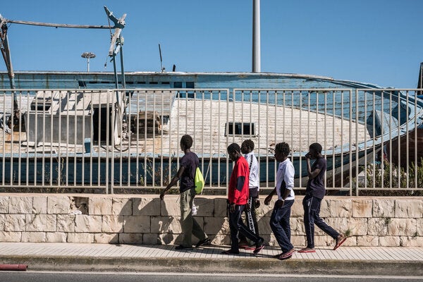 De jeunes Gambiens passent près d'un bateau de contrebande dans un cimetière de navires, dans le port près du centre de réception pour les demandeurs d'asile qui sert également de centre d'hébergement pour les mineurs non-accompagnés à Pozzallo, en Sicile, en Italie (Mai 2016).