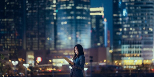 Pretty office lady is using digital tablet, standing against illuminated highrise corporate buildings at night time.