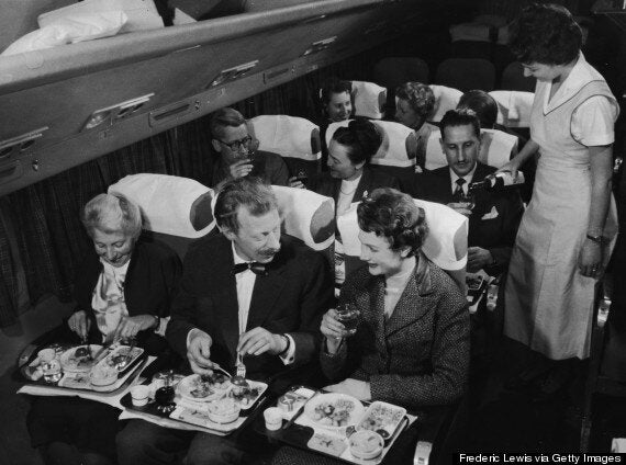 Interior view of a commercial passenger plane shows, in the foreground, a couple as they enjoy their meal next to a smiling elderly woman, while behind them, a flight attendant pours a glass of wine for a man who sits next to a couple who toast each other with full glasses, 1950s. (Photo by Frederic Lewis/Getty Images)