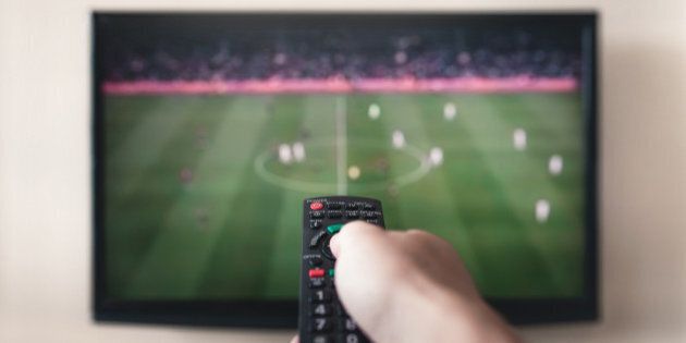 Human hand holding remote control with soccer channel on the television screen.