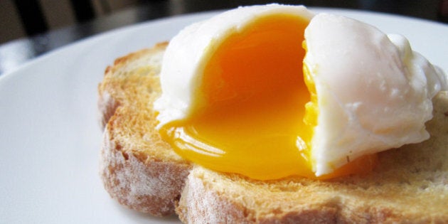 A single poached egg on artisan hand sliced toasted bread. The egg is split to reveal a runny yolk. White dinner plate, non-descript dark wood table and chair just about visible in the background.