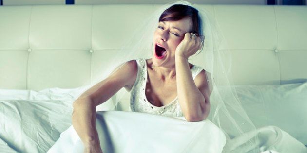 A young woman sitting on a bed wearing a wedding dress and veil yawning.