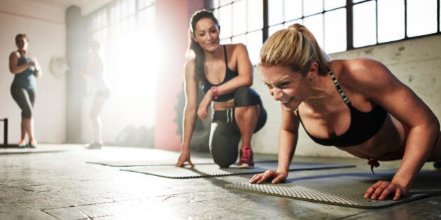 Healthy woman fitness training, doing press-ups with determined expression in urban industrial gym. Friend is giving encouragement beside her whilst other females workout in background.