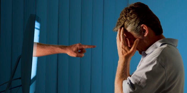 Severely distraught young man sitting in front of a computer with a judgmental hand pointing at him from within the computer monitor which shows the man being either computer bullying bullied or Facebook social media stalking stalked.