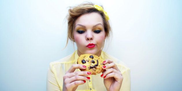 Young woman with ginger hair wearing a yellow blouse and colourful make-up staring at a smiling chocolate chip cookie.