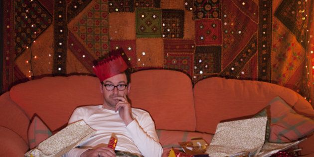 A man wearing a paper hat and surrounded by Christmas wrapping paper, slumps alone on a sofa in front of a TV.