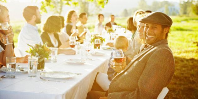 Man sitting at end of banquet table outside in field laughing holding glass of wine