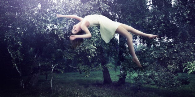 A female wearing a white dress is floating in a forest. There is a strong light source just above her.