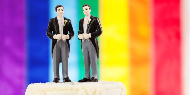 Subject: Same sex marriage wedding cake with two male groom figurine cake toppers and rainbow flag in background.