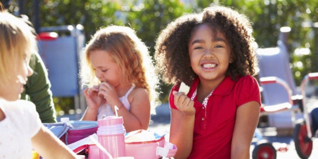 Elementary Pupils Sitting At Table Eating Lunch Smiling To Camera Holding Snack