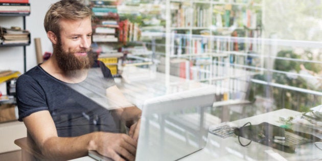 Young bearded man sitting on desk in his home office and working on laptop.