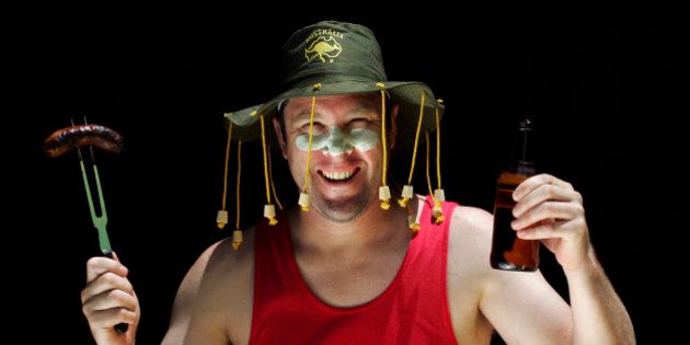 An Aussie man in a cork hat smiles at the camera holding up a sausage on a fork in one hand and a beer in the other