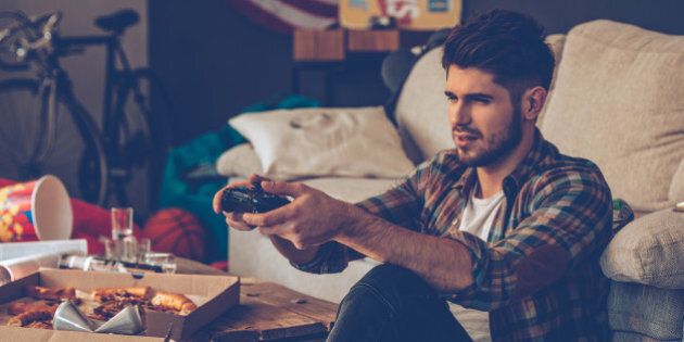 Frustrated young man holding joystick while sitting on the floor in messy room after party