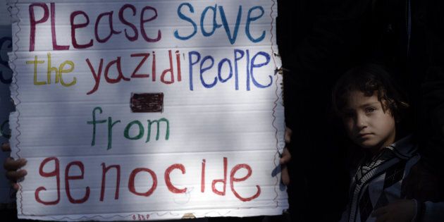 A migrant child stands next to a placard reading 'Please save the Yazidi people from genocide' at the Moria detention center during a visit of Pope Francis in Mytilene, on the Greek island of Lesbos, on April 16, 2016.'We are all migrants,' Pope Francis said on April 16 on a visit to the Greek island of Lesbos to give hope to thousands facing deportation after risking their lives to reach Europe. The pontiff's visit came amid continuing criticism of a deal reached last month to end Europe's refugee crisis by sending all irregular migrants who land in Greece back to Turkey. / AFP / ARIS MESSINIS (Photo credit should read ARIS MESSINIS/AFP/Getty Images)