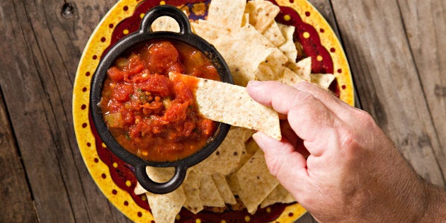 An overhead close up shot of a hand dipping a corn chip into a black cast iron salsa bowl sitting on a festive plate full of chips. Shot on an old grungy wooden table.