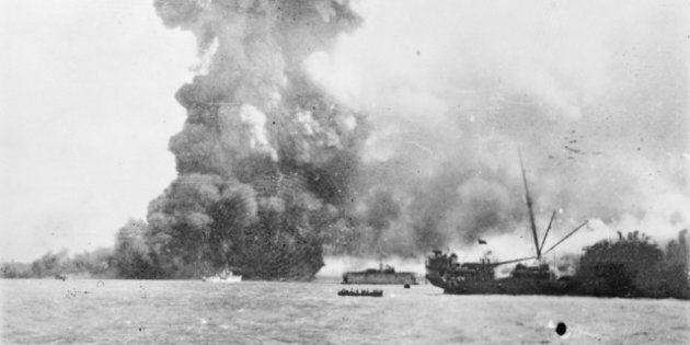 The SS Neptuna explodes at Stokes Hill Wharf, sending a column of debris and smoke hundreds of metres into the air. Directly in front of the explosion is the small Patrol Craft HMAS Vigilant which is undertaking rescue work. In the centre background the floating dry dock holding the Corvette, HMAS Katoomba is visible. In the centre foreground a lifeboat pulls away from the damaged SS Zealandia (right) which was dive bombed and later sunk.
