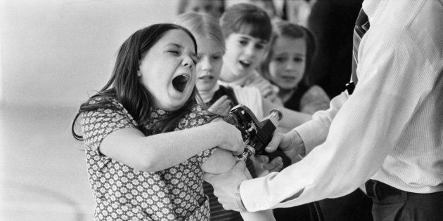 Classmates look on as a girl overreacts to anti-measles vaccination in the 1970s.