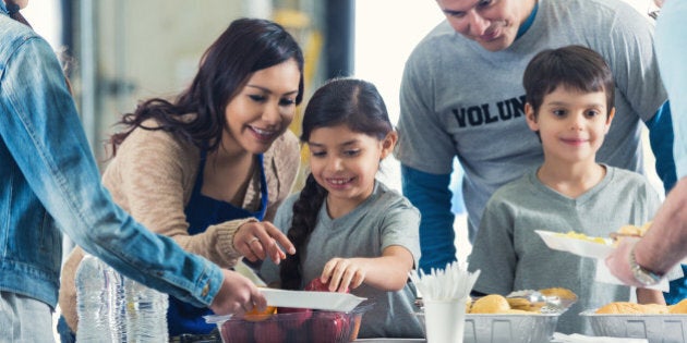 Young Hispanic family is serving healthy meals to people in charity food bank soup kitchen. Mother and father are instructing elementary age little boy and little girl as they serve food to senior man and preteen girl.