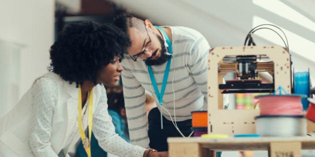 Creative Multi-Ethnic start-up business team working by 3D printer. Standing and looking at 3d printer and printed object. Male with headphones is looking at printed object in 3D printer. They are wearing name tags.