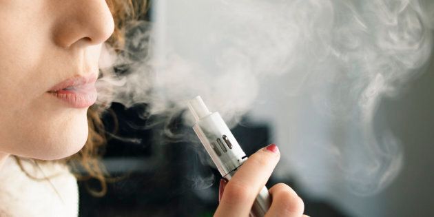 Close-up of a young woman exhaling smoke vapour from an electronic cigarette, holding the device in her hand. Shot at home, indoors.