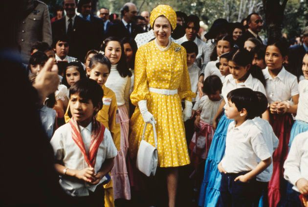 Queen Elizabeth II sporting a turban hat that matches her yellow and white gown, with a group of local children during a state visit to Mexico, February-March 1975.
